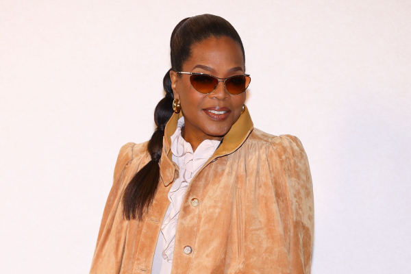 Oprah appears at a fashion show wearing a suede jacket blouse and bubble ponytail with sunglasses.